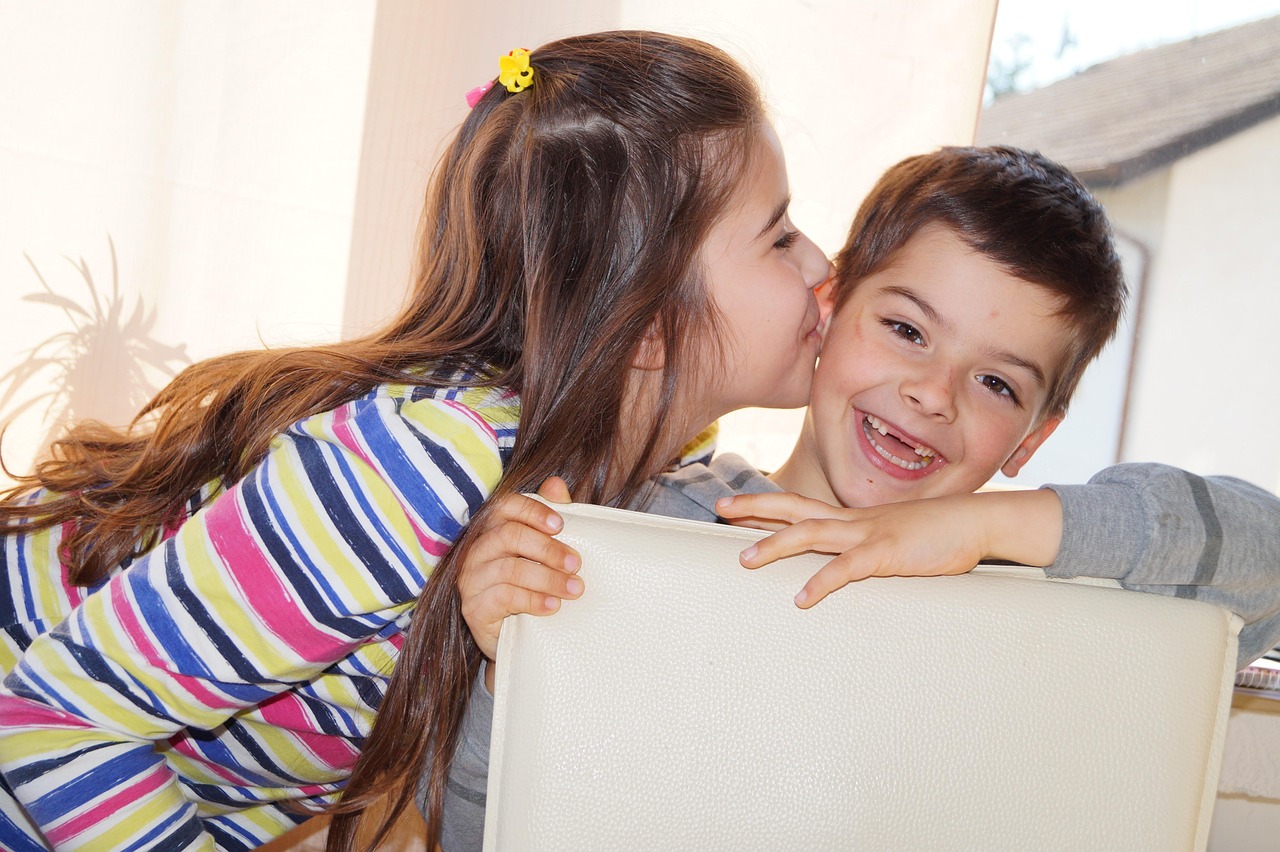 Younger sister with long brown hair kisses her brother on the cheek indoors, both enjoying a playful moment in colorful clothing.